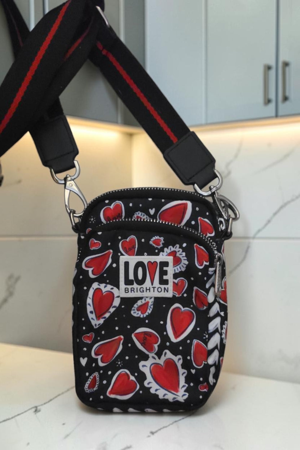 The Gettin Place Brighton crossbody bag in black with red heart print, featuring a LOVE BRIGHTON patch and strap.