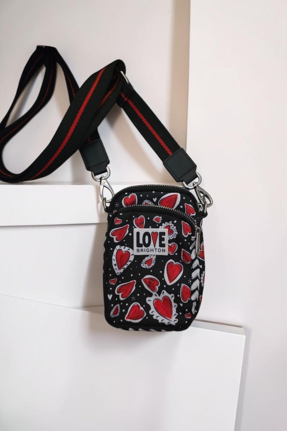 The Gettin Place black crossbody bag with red heart print, LOVE BRIGHTON patch, and adjustable red-striped strap.