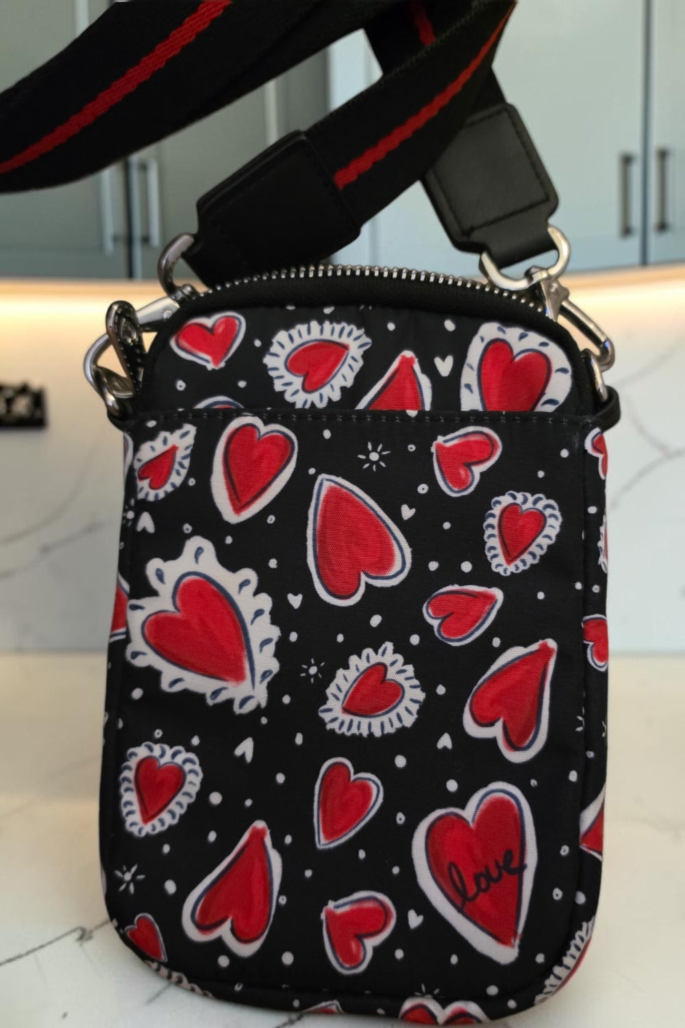 The Gettin Place black crossbody bag with red heart print, featuring a branded LOVE patch and adjustable strap.