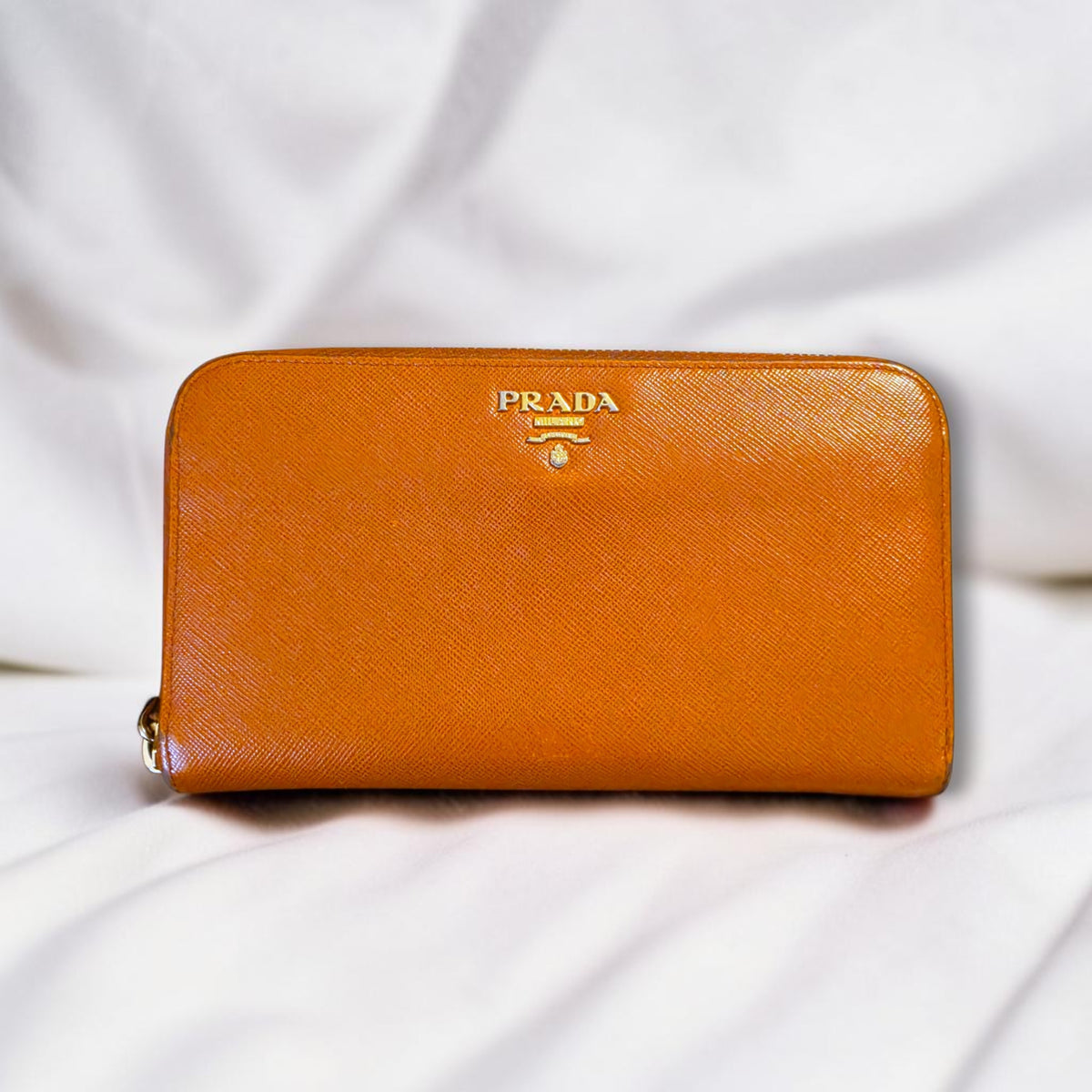 The Gettin Place orange Saffiano leather Prada zip-around wallet with gold-tone logo plaque on a white fabric background